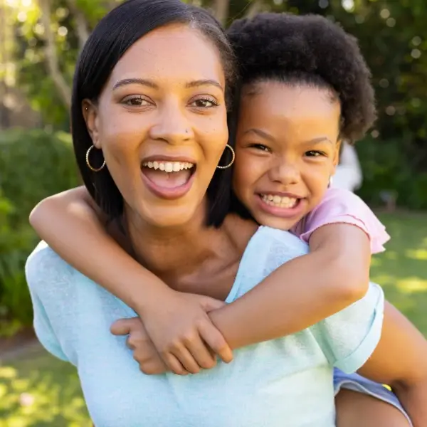 A smiling mother gives her young daughter a piggyback ride outdoors, both laughing together in a green backyard setting.