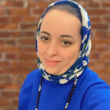 Portrait of Raja Alabyadh smiling in a blue floral hijab and blue top against a brick wall background.